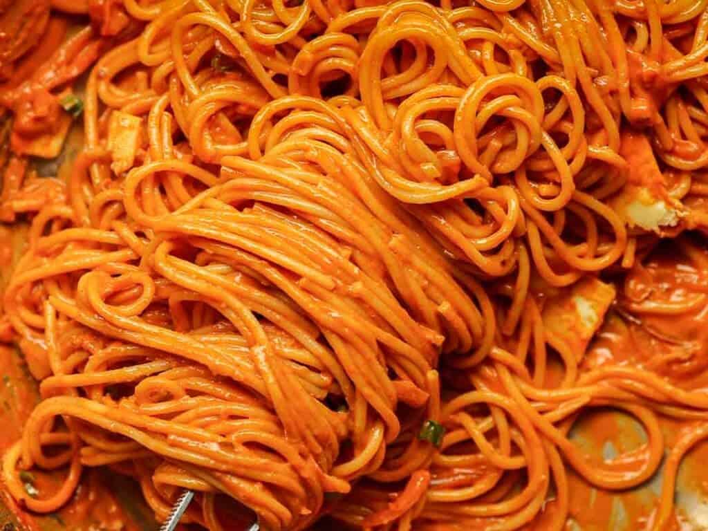 A close-up of spaghetti covered in a thick, red sauce. The noodles are tangled and saturated with the vibrant sauce, giving the dish a rich appearance. Small bits of herbs and spices are visible throughout.